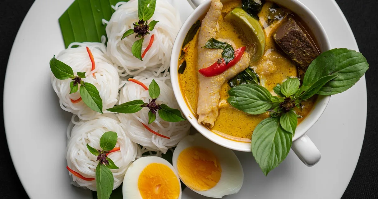 Prato com macarrão de arroz, ovos cozidos e uma tigela de curry com frango e manjericão
